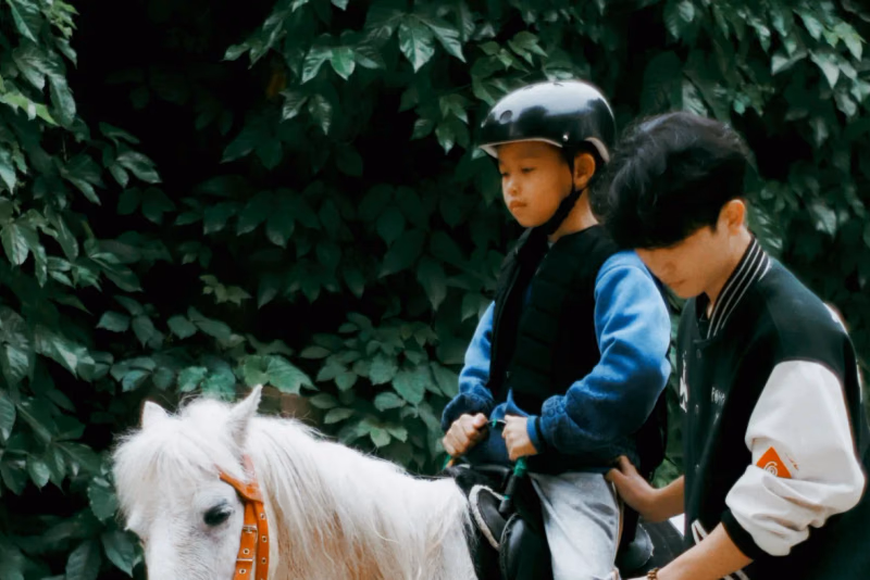 Kind mit Reithelm und Sicherheitsweste sitzt auf einem weißen Pony, ein Erwachsener führt und sichert es